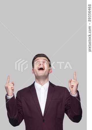 Portrait of surprised handsome young man in violet suit and white shirt, standing, looking top and pointing at copy space on top with amazed face. indoor studio shot, isolated on grey background 89356468