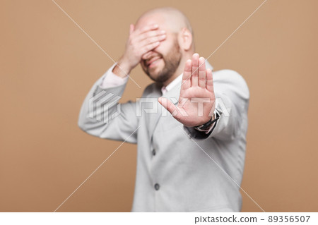 Stop it, I dont want to see it. Portrait of confused middle aged bald bearded businessman in classic light gray suit standing with stop gesture, covered his eyes. studio shot on brown background. 89356507