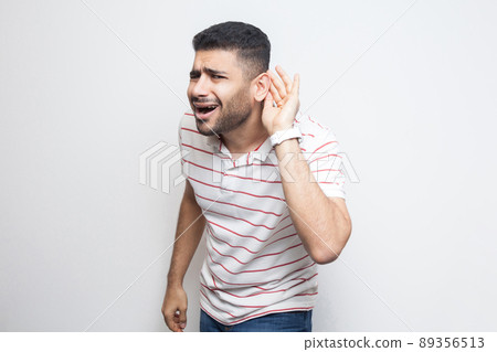What? I can not hear you. Portrait of attentive handsome bearded young man in striped t-shirt standing with hand on ear and want to hear something. indoor studio shot, isolated on white background. What? I can not hear you. Portrait of attentive handsome bearded young man in striped t-shirt standing with hand on ear and want to hear something. indoor studio shot, isolated on white background. 89356513