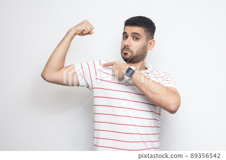 I am strong. Portrait of proud handsome bearded young man in striped t-shirt standing, pointing at his bicep and looking at camera with satisfied face. indoor studio shot, isolated on white background I am strong. Portrait of proud handsome bearded young man in striped t-shirt standing, pointing at his bicep and looking at camera with satisfied face. indoor studio shot, isolated on white background 89356542