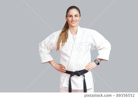 Happy cheerful beautiful blonde young athletic woman in white kimono, black belt standing, holdind hands on waist, toothy smiling and looking at camera. Indoor studio shot, isolated on grey background 89356552
