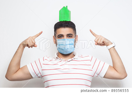 Stay at home, stay safe. Positive man with surgical medical mask showing green paper small house on head, propose to be at home on quarantine time. Indoor, isolated, studio shot, white background 89356555