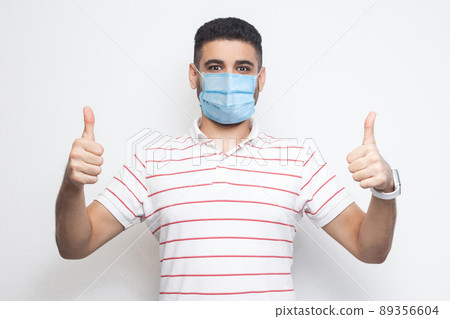 I like this. I am safe. Portrait of satisfied happy young man with surgical medical mask in striped t-shirt standing, thumbs up and looking at camera. indoor studio shot, isolated on white background. I like this. I am safe. Portrait of satisfied happy young man with surgical medical mask in striped t-shirt standing, thumbs up and looking at camera. indoor studio shot, isolated on white background. 89356604