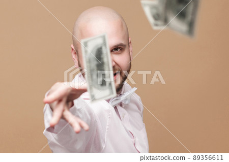 Portrait of happy handsome middle aged bald bearded businessman in white shirt standing, throw out money and looking at camera with proud rich face. studio shot, isolated on light brown background. 89356611