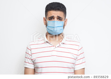 Portrait of sad upset or bored man with surgical medical mask in striped t-shirt standing and looking at camera with dissatisfied sadness face. indoor studio shot, isolated on white background. 89356629
