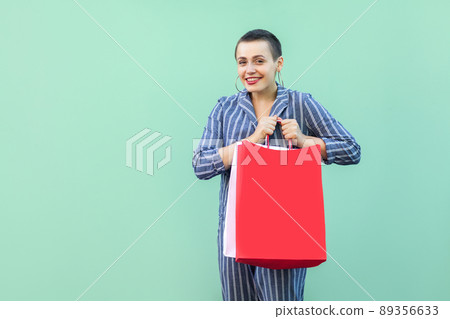 Portrait of happy beautiful with short hair young woman in striped suit standing, holding paper package, looking at camera with toothy smile. Indoor, isolated, studio shot, green background Portrait of happy beautiful with short hair young woman in striped suit standing, holding paper package, looking at camera with toothy smile. Indoor, isolated, studio shot, green background 89356633