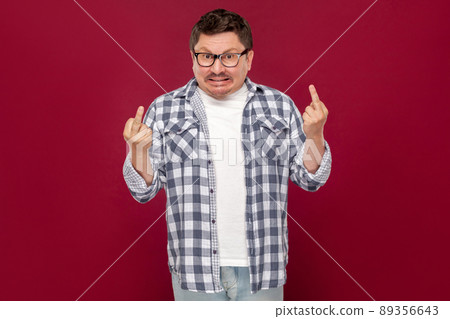 Portrait of angry middle aged business man in casual checkered shirt and eyeglasses standing and looking at camera with middle finger fuck sign. indoor studio shot, isolated on dark red background. 89356643
