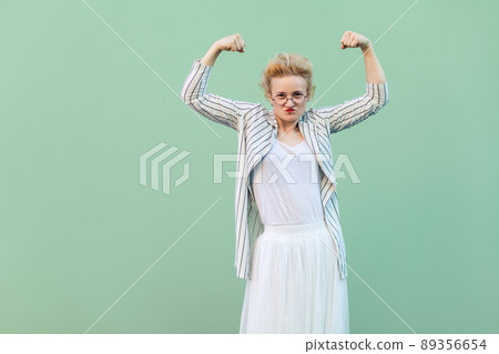 independence woman. Portrait of strong proud young blonde woman in white striped blouse with eyeglasses standing with raised arms and looking at camera. studio shot isolated on light green background 89356654