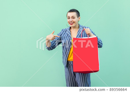 Portrait of satisfied beautiful with short hair young woman in striped suit standing, holding shopping red bag and pointing finger to it. Indoor, isolated, studio shot, green background Portrait of satisfied beautiful with short hair young woman in striped suit standing, holding shopping red bag and pointing finger to it. Indoor, isolated, studio shot, green background 89356664
