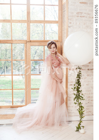 Full length portrait of sensual dreaming beautiful pregnant young woman in pink dress standing near big natural window and touching her neck with hand. Last months of pregnancy, studio shot, isolated Full length portrait of sensual dreaming beautiful pregnant young woman in pink dress standing near big natural window and touching her neck with hand. Last months of pregnancy, studio shot, isolated 89356676
