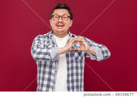 Portrait of happy handsome middle aged man in heckered shirt, eyeglasses standing with heart shape gesture hands and looking at camera with toothy smile. studio shot, isolated on dark red background Portrait of happy handsome middle aged man in heckered shirt, eyeglasses standing with heart shape gesture hands and looking at camera with toothy smile. studio shot, isolated on dark red background 89356677