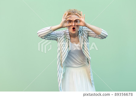 Portrait of surprised young blonde woman in white shirt, skirt, and striped blouse with eyeglasses standing with binoculars hands on eyes. indoor studio shot isolated on light green background. Portrait of surprised young blonde woman in white shirt, skirt, and striped blouse with eyeglasses standing with binoculars hands on eyes. indoor studio shot isolated on light green background. 89356678