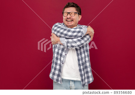 Portrait of happy satisfied handsome middle aged business man in casual checkered shirt, eyeglasses standing, hugging himself smiling and enjoying. indoor studio shot, isolated on dark red background. Portrait of happy satisfied handsome middle aged business man in casual checkered shirt, eyeglasses standing, hugging himself smiling and enjoying. indoor studio shot, isolated on dark red background. 89356680
