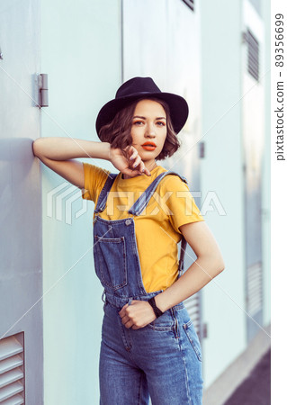 young woman in blue denim overalls and yellow tshirt with black hat sensual looking at camera while posing near industrial building . outdoor shot in the summertime young woman in blue denim overalls and yellow tshirt with black hat sensual looking at camera while posing near industrial building . outdoor shot in the summertime 89356699