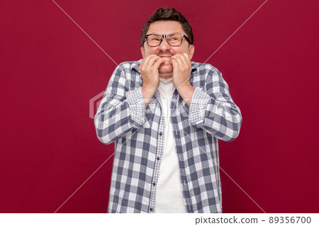 Portrait of worried nervous or stressed middle aged business man in casual checkered shirt and eyeglasses standing, bitting his nails and looking away. studio shot, isolated on dark red background. 89356700