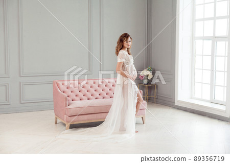 Full length portrait of beautiful pregnant young woman in white dress peignoir standing near pink sofa, holding with love her belly, posing like model. Last months of pregnancy, studio shot, isolated Full length portrait of beautiful pregnant young woman in white dress peignoir standing near pink sofa, holding with love her belly, posing like model. Last months of pregnancy, studio shot, isolated 89356719