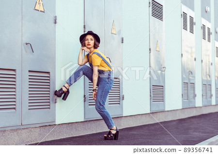 young woman in blue denim overalls and yellow tshirt with black hat sensual looking at camera while posing near industrial building . outdoor shot in the summertime 89356741
