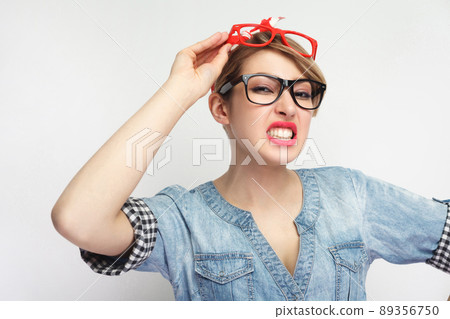 Anger sexual young woman in casual blue denim shirt with makeup standing, wearing lots of colorful spectacles at the same time, clench one's teeth . indoor studio shot, isolated on white background 89356750