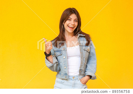 Portrait of happy satisfied beautiful brunette young woman with makeup in denim casual style standing, thumbs up and looking at camera excited. indoor studio shot, isolated on yellow background. Portrait of happy satisfied beautiful brunette young woman with makeup in denim casual style standing, thumbs up and looking at camera excited. indoor studio shot, isolated on yellow background. 89356758