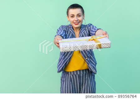 Portrait of friendly beautiful with short hair young woman in striped suit standing, giving you, congratulation you with present box, looking at camera. Indoor, isolated, studio shot, green background 89356764