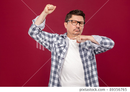 I am strong. Portrait of proud handsome middle aged business man in casual checkered shirt and eyeglasses standing and pointing at his biceps. indoor studio shot, isolated on dark red background. I am strong. Portrait of proud handsome middle aged business man in casual checkered shirt and eyeglasses standing and pointing at his biceps. indoor studio shot, isolated on dark red background. 89356765