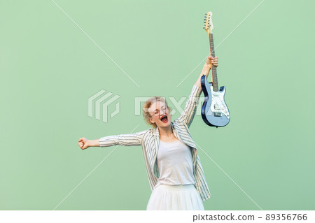 Portrait of surprised joyful young blonde woman in white striped blouse with eyeglasses holding electric guitar standing and rejoicing her victory. indoor studio shot isolated, light green background. Portrait of surprised joyful young blonde woman in white striped blouse with eyeglasses holding electric guitar standing and rejoicing her victory. indoor studio shot isolated, light green background. 89356766