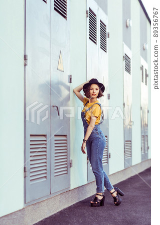 young woman in blue denim overalls and yellow tshirt with black hat sensual looking at camera while posing near industrial building . outdoor shot in the summertime 89356767