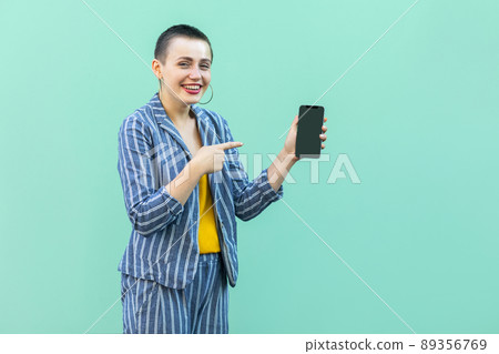 Happy beautiful with short hair young woman in striped suit standing, holding phone and pointing finger to it, toothy smile and looking at camera. Indoor, isolated, studio shot, green background 89356769