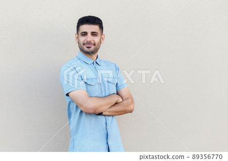 Portrait of confident successful handsome young bearded businessman in blue shirt standing, crossed arms and looking at camera with smile. indoor studio shot isolated on light beige wall background. 89356770