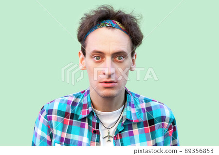 Closeup portrait of confused young man in casual blue checkered shirt and headband standing and looking at camera with shocked serious face. indoor studio shot, isolated on light green background. 89356853