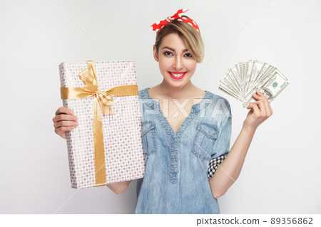 Portrait of beautiful young woman in casual blue denim shirt with makeup and red headband standing, showing gift box and fan dollars with toothy smile. indoor studio shot, isolated on white background 89356862