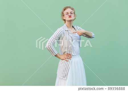 Portrait of proud young woman in white striped blouse with eyeglasses standing with hand on waist, pointing herself and looking at camera. studio shot isolated on light green background. Portrait of proud young woman in white striped blouse with eyeglasses standing with hand on waist, pointing herself and looking at camera. studio shot isolated on light green background. 89356888