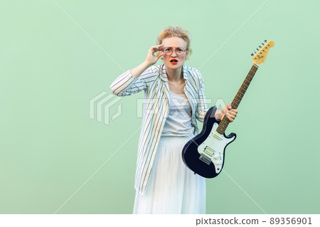Portrait of young blonde woman in white shirt and striped blouse, electric guitar standing, holding eyeglasses and looking attentive at camera. indoor studio shot isolated on light green background. 89356901