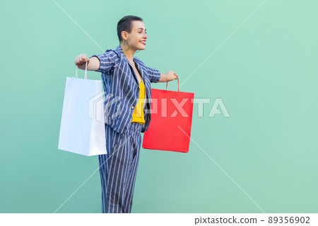 Side view of satisfied with short hair young woman in striped suit standing, holding shopping bags with raised arms, toothy smile and looking at camera. Indoor, isolated, studio shot, green background Side view of satisfied with short hair young woman in striped suit standing, holding shopping bags with raised arms, toothy smile and looking at camera. Indoor, isolated, studio shot, green background 89356902