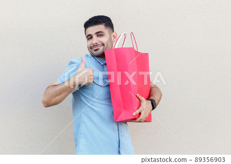 Amazing gift. Portrait of satisfied young bearded man in blue shirt standing, holding red shopping bag, thumbs up, looking at camera with smile. studio shot isolated on light beige wall background. Amazing gift. Portrait of satisfied young bearded man in blue shirt standing, holding red shopping bag, thumbs up, looking at camera with smile. studio shot isolated on light beige wall background. 89356903