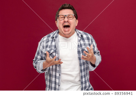 Portrait of handsome middle aged business man in casual checkered shirt and eyeglasses standing, raised arms, looking at camera and screaming. indoor studio shot, isolated on dark red background. 89356941