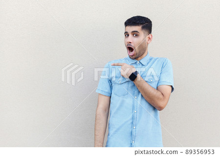 Portrait of shocked handsome young bearded man in blue shirt standing, looking at camera with surprised face and pointing at copyspace. indoor studio shot isolated on light beige wall background. 89356953