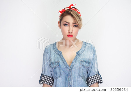 Portrait of sad beautiful young woman in casual blue denim shirt with makeup and red headband standing and looking at camera with unhappy face. indoor studio shot, isolated on white background. Portrait of sad beautiful young woman in casual blue denim shirt with makeup and red headband standing and looking at camera with unhappy face. indoor studio shot, isolated on white background. 89356984
