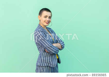 Portrait of happy handsome beautiful short hair young stylish woman in casual striped suit standing, crossed hands, looking at camera with toothy smile. studio shot isolated on light green background. Portrait of happy handsome beautiful short hair young stylish woman in casual striped suit standing, crossed hands, looking at camera with toothy smile. studio shot isolated on light green background. 89356985