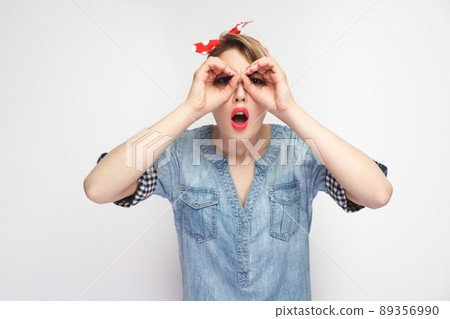 Portrait of surprised beautiful young woman in casual blue denim shirt with makeup and red headband standing with binoculars hands gesture and looking. indoor studio shot, isolated on white background Portrait of surprised beautiful young woman in casual blue denim shirt with makeup and red headband standing with binoculars hands gesture and looking. indoor studio shot, isolated on white background 89356990