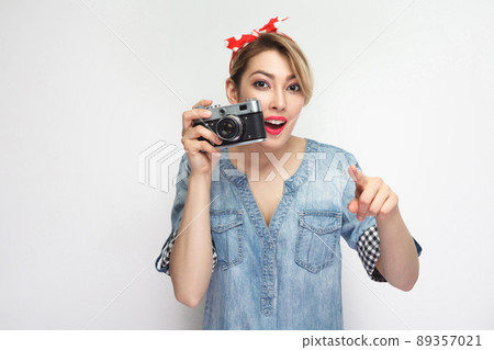 Want to make photoshout. Young blogger girl in casual denim shirt with makeup, red headband standing, holding retro camera and pointing finger to you. indoor studio shot, isolated on white background Want to make photoshout. Young blogger girl in casual denim shirt with makeup, red headband standing, holding retro camera and pointing finger to you. indoor studio shot, isolated on white background 89357021