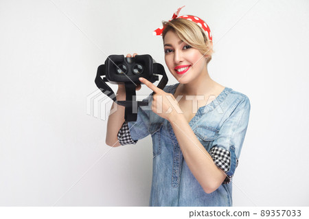 Happy young woman in casual blue denim shirt, red headband holding virtual reality googles VR and pointing finger to back view, toothy smile. indoor studio shot, isolated on white background 89357033