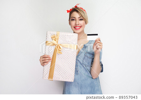 Portrait of beautiful young woman in casual blue denim shirt with makeup and red headband standing, showing gift box and credit card with toothy smile. indoor studio shot, isolated on white background 89357043