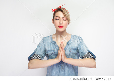 Portrait of calm beautiful young woman in casual blue denim shirt with makeup and red headband standing with palm hands and meditating in yoga namaste pose. studio shot, isolated on white background. 89357048