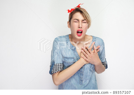 Heart attack. Portrait of young woman in casual blue denim shirt with makeup and red headband standing screaming and holding her painful heart. indoor studio shot, isolated on white background. Heart attack. Portrait of young woman in casual blue denim shirt with makeup and red headband standing screaming and holding her painful heart. indoor studio shot, isolated on white background. 89357107