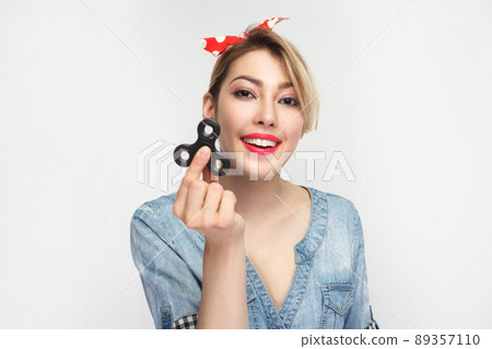 Portrait of beautiful happy young woman in casual blue denim shirt with makeup and red headband standing and holding hand spinner, toothy smile. indoor studio shot, isolated on white background. 89357110