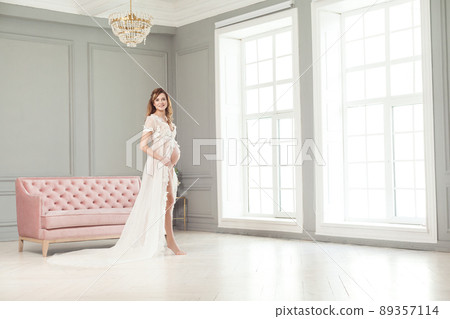 Full length portrait of beautiful pregnant young woman in white peignoir standing near pink sofa, holding with love her belly. Last months of pregnancy, studio shot, looking at camera, toothy smile Full length portrait of beautiful pregnant young woman in white peignoir standing near pink sofa, holding with love her belly. Last months of pregnancy, studio shot, looking at camera, toothy smile 89357114
