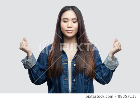 Portrait of calm beautiful brunette asian young woman in casual blue denim jacket with makeup standing with raised arms and yoga meditating. indoor studio shot, isolated on light grey background. 89357115