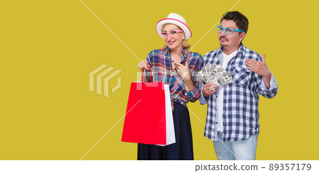 Happy family after shopping, adult man and woman in casual checkered shirt standing together,wife holding paper bag with toothy smile,husband fan of money like big sponsor.Indoor, isolated,studio shot Happy family after shopping, adult man and woman in casual checkered shirt standing together,wife holding paper bag with toothy smile,husband fan of money like big sponsor.Indoor, isolated,studio shot 89357179