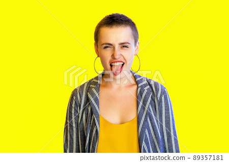 Portrait of funny crazy young short hair beautiful woman in yellow shirt and striped suit standing and looking at camera with tongue out. indoor studio shot isolated on yellow background. 89357181